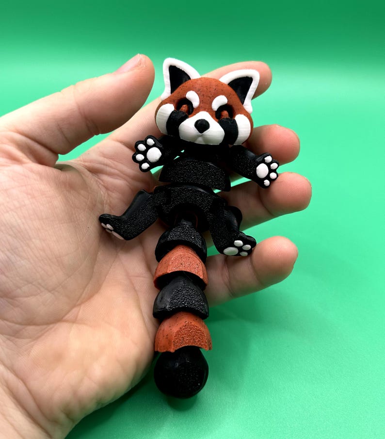 May include: A 3D-printed red panda figurine with a segmented body. The panda has a red and white head with black accents, black limbs, and a segmented tail in alternating black and red-orange colors. The figurine is held against a green background.