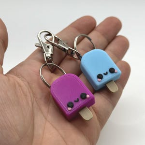 Pu&ograve; includere: Due portachiavi a forma di ghiacciolo, uno viola e uno azzurro, ciascuno con una faccina sorridente e un bastoncino di legno. I portachiavi sono attaccati a moschettoni e anelli in metallo argentato.