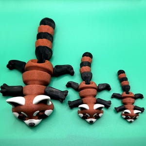 May include: Three red panda figurines in varying sizes. Each has a brown body, black limbs, and a striped black and brown tail. The faces are brown with white markings. The figurines are arranged on a solid green background.