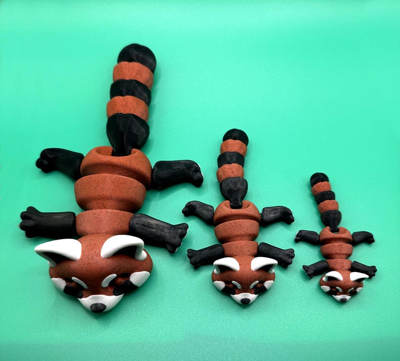 May include: Three red panda figurines in varying sizes. Each has a brown body, black limbs, and a striped black and brown tail. The faces are brown with white markings. The figurines are arranged on a solid green background.