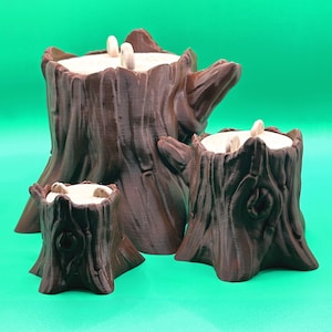 May include: Three brown tree stump-shaped candle holders of varying sizes. Each has a textured, bark-like exterior and a light-colored interior with three white candles. The candle holders are arranged against a solid green background.