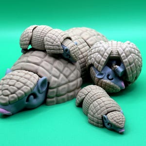 May include: A collection of whimsical, stylized armadillo figurines in various sizes. The armadillos have tan, segmented shells and blue-grey faces with closed eyes and pink tongues. They are arranged on a solid green background.