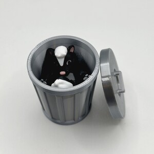 May include: A small, gray plastic trash can with a lid. A black and white skunk figurine is inside the trash can.