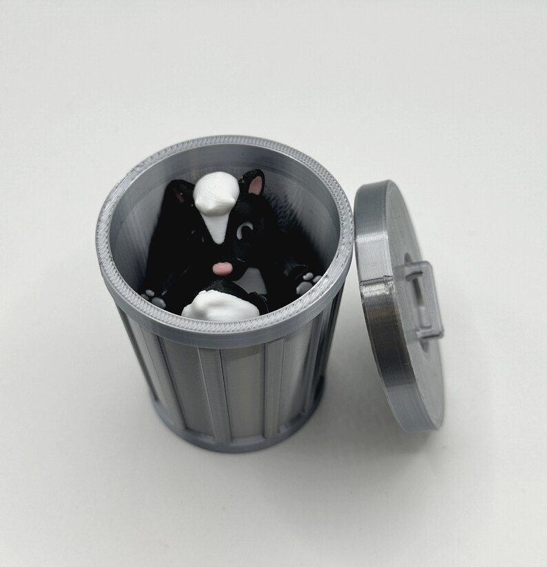 May include: A small, gray plastic trash can with a lid. A black and white skunk figurine is inside the trash can.