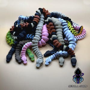 May include: A collection of articulated, segmented snake figurines in various colours including black, pink, grey, blue, green, and brown. Each snake features a detailed head and a flexible, jointed body. The text NEBULA SQUID appears with a logo.