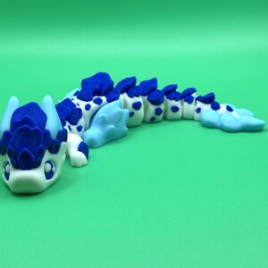 May include: A handcrafted, whimsical dragon figurine in shades of blue and white. The dragon features a detailed head with a crown-like structure and a segmented body with blue spots. The figurine is set against a solid green background.