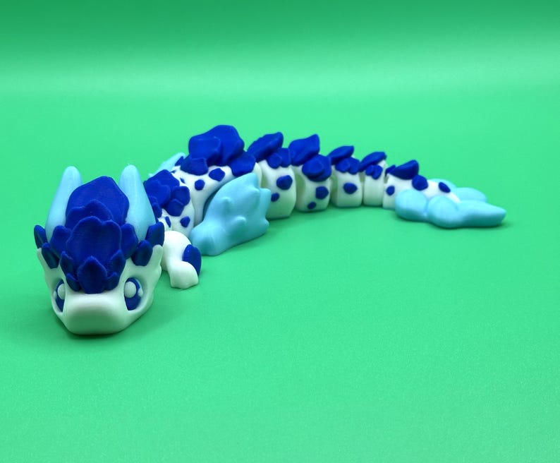May include: A handcrafted, whimsical dragon figurine in shades of blue and white. The dragon features a detailed head with a crown-like structure and a segmented body with blue spots. The figurine is set against a solid green background.