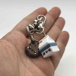 Pu&ograve; includere: Un portachiavi con un biscotto con gocce di cioccolato e un ciondolo a forma di cartone del latte. Il biscotto &egrave; marrone con gocce marroni scure e guance rosa. Il cartone del latte &egrave; bianco con una fascia blu e guance rosa.