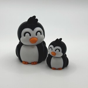 Op de afbeelding: Twee pingu&iuml;nbeeldjes. De pingu&iuml;ns zijn zwart en wit met oranje snavels en voeten. De grotere pingu&iuml;n is ongeveer 7,6 cm hoog en de kleinere is ongeveer 5 cm hoog. Ze hebben een cartoonachtig ontwerp met een glimlachende uitdrukking.
