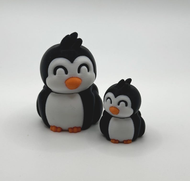 Op de afbeelding: Twee pingu&iuml;nbeeldjes. De pingu&iuml;ns zijn zwart en wit met oranje snavels en voeten. De grotere pingu&iuml;n is ongeveer 7,6 cm hoog en de kleinere is ongeveer 5 cm hoog. Ze hebben een cartoonachtig ontwerp met een glimlachende uitdrukking.
