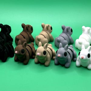 May include: A collection of ten small, stylized bunny figurines in various colors, including black, brown, tan, gray, and white. Each bunny has large black eyes and pink noses, set against a vibrant green background. These decorative figurines are ideal for Easter.