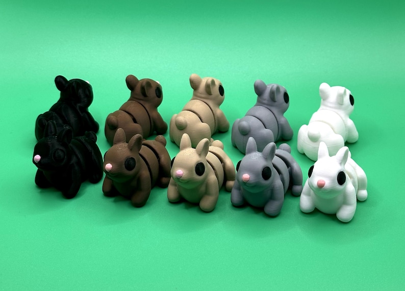 May include: A collection of ten small, stylized bunny figurines in various colors, including black, brown, tan, gray, and white. Each bunny has large black eyes and pink noses, set against a vibrant green background. These decorative figurines are ideal for Easter.