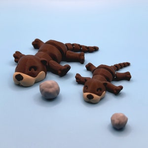 Op de afbeelding: Twee bruine, gearticuleerde otter-figuurtjes met beschilderde details, liggend op een lichtblauw oppervlak. Twee kleine grijze stenen liggen ook op het oppervlak.