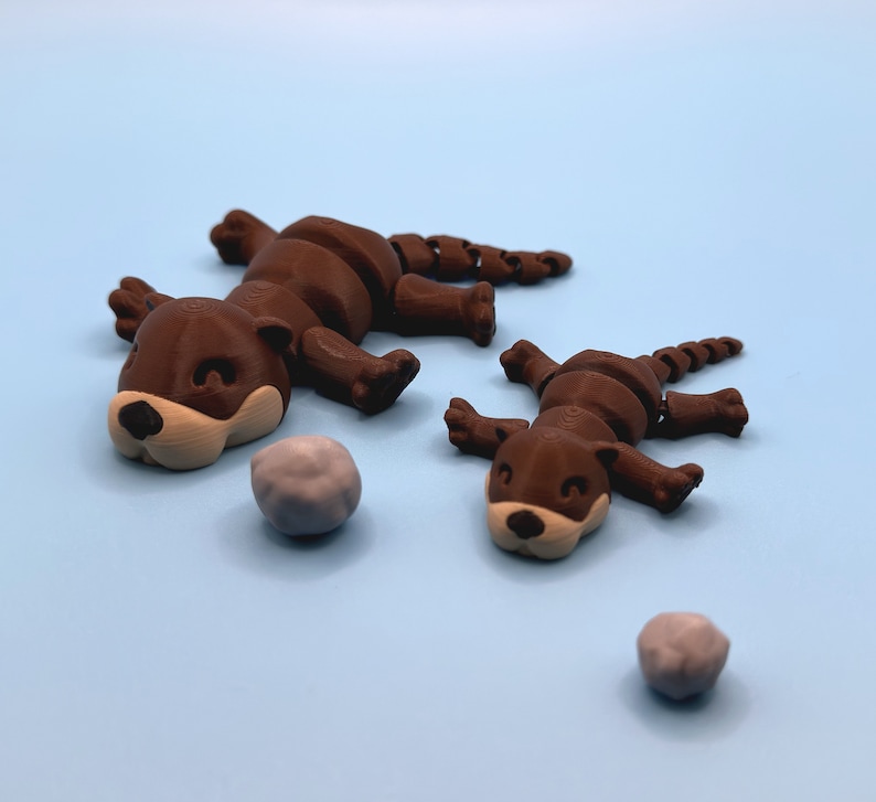 Op de afbeelding: Twee bruine, gearticuleerde otter-figuurtjes met beschilderde details, liggend op een lichtblauw oppervlak. Twee kleine grijze stenen liggen ook op het oppervlak.