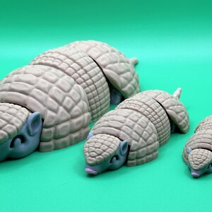 May include: Three armadillo figurines in varying sizes, arranged on a solid green background. The armadillos have a light tan, segmented shell and blue-gray heads with small ears and closed eyes. The smallest armadillo is about 2 inches long.