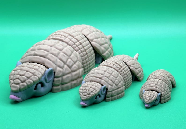 May include: Three armadillo figurines in varying sizes, arranged on a solid green background. The armadillos have a light tan, segmented shell and blue-gray heads with small ears and closed eyes. The smallest armadillo is about 2 inches long.