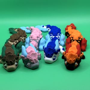 May include: A collection of colorful, handcrafted dragon figurines in various colors, including green, brown, pink, blue, and orange. Each dragon has a unique design and is set against a solid green background.