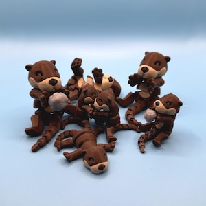 Op de afbeelding: Een groep van zes bruine, gearticuleerde otter-figuurtjes, waarvan sommige kleine grijze stenen vasthouden, zijn speels geposeerd op een lichtblauwe achtergrond.