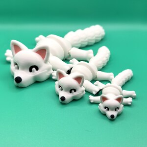 May include: Three white articulated fox figurines in different sizes. The largest fox is lying down with its head turned to the side. The medium-sized fox is lying down with its head turned to the side. The smallest fox is lying down with its head turned to the side.