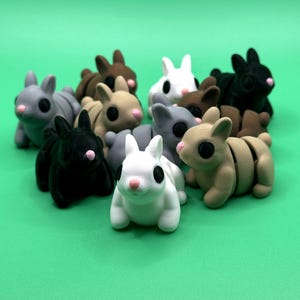 May include: A collection of small, 3D-printed bunny figurines in various colors, including black, white, gray, and brown. Each bunny has large black eyes and a pink nose. The bunnies are arranged on a solid green background.