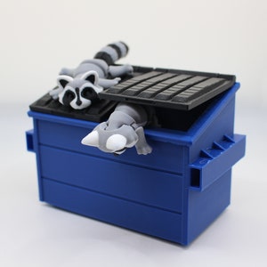 May include: A blue plastic recycling bin with a raccoon figurine peeking out of the top. The raccoon is gray and white with black eyes and a black nose.