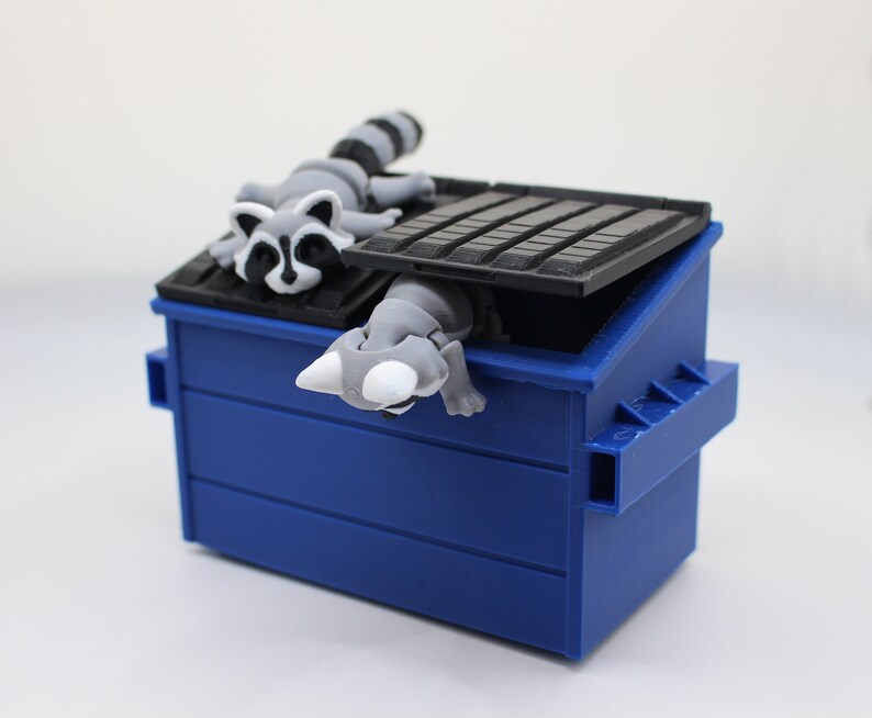 May include: A blue plastic recycling bin with a raccoon figurine peeking out of the top. The raccoon is gray and white with black eyes and a black nose.