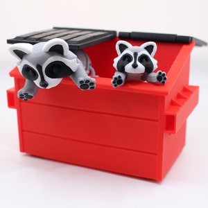 May include: Two raccoons peeking out of a red plastic trash can with a black lid. The raccoons are white and gray with black eyes and noses.