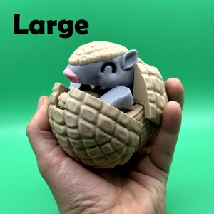 May include: A tan and gray 3D-printed armadillo toy with a smiling face and a pink nose. The armadillo is partially enclosed in a segmented shell. The word "Large" is printed in black at the top.