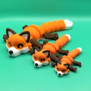 May include: Three 3D printed orange and brown fox figurines of varying sizes. The largest fox is lying down with its head resting on the surface. The two smaller foxes are also lying down with their heads resting on the surface.