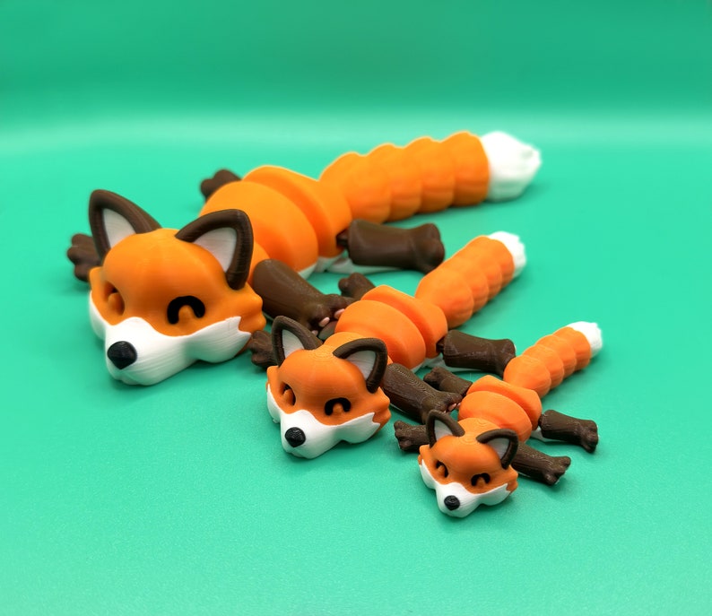 May include: Three 3D printed orange and brown fox figurines of varying sizes. The largest fox is lying down with its head resting on the surface. The two smaller foxes are also lying down with their heads resting on the surface.