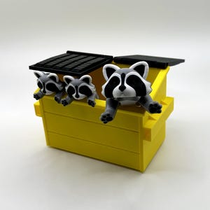 May include: A yellow toy dumpster with a black lid and three gray and white raccoon figurines peeking over the edge.