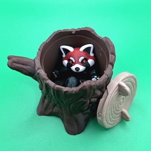 May include: A brown tree stump-shaped container with a red panda figurine inside. The panda has a red head with white markings and is wearing a black outfit. The container has a removable lid, and the background is a solid green.