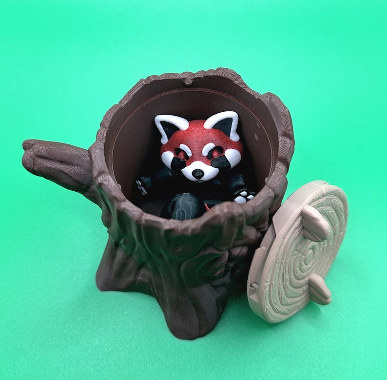 May include: A brown tree stump-shaped container with a red panda figurine inside. The panda has a red head with white markings and is wearing a black outfit. The container has a removable lid, and the background is a solid green.