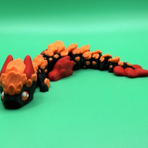 May include: A handcrafted, stylized dragon figurine in black, orange, and red. The dragon has a detailed head with large eyes and a segmented body. The figurine is set against a solid green background.
