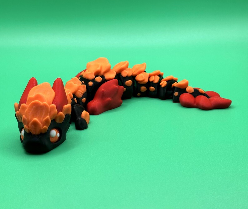May include: A handcrafted, stylized dragon figurine in black, orange, and red. The dragon has a detailed head with large eyes and a segmented body. The figurine is set against a solid green background.