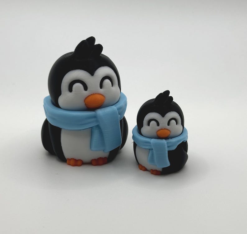 Op de afbeelding: Twee pingu&iuml;nbeeldjes, een grote en een kleine, worden getoond. De pingu&iuml;ns zijn zwart en wit met oranje snavels en voeten. Elk draagt een lichtblauwe sjaal. De pingu&iuml;ns hebben een vrolijke uitdrukking.