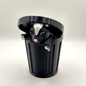May include: A black and white skunk figurine is peeking out of a black trash can with a lid. The skunk has a white stripe down its back and pink nose.