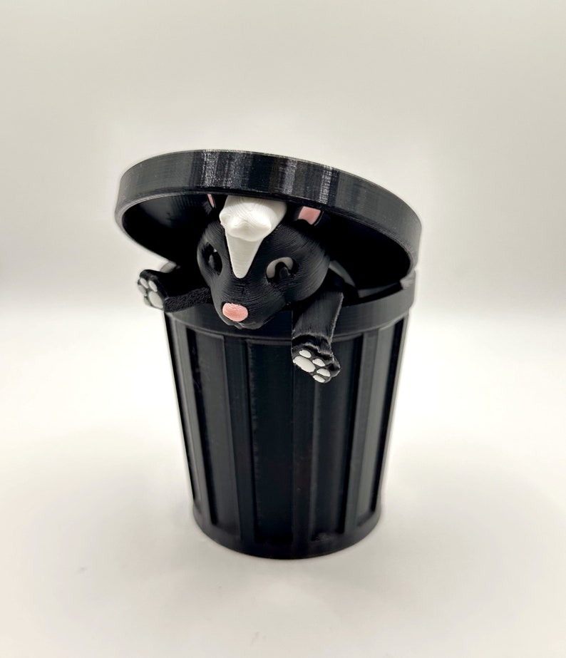 May include: A black and white skunk figurine is peeking out of a black trash can with a lid. The skunk has a white stripe down its back and pink nose.
