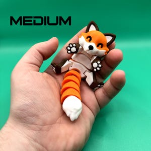 May include: A small, orange and white fox-shaped toy with a segmented tail. The toy is made of plastic and has a friendly expression. The toy is being held in a person's hand.