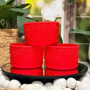 May include: Three red, cylindrical planters with a woven geometric pattern. The planters are arranged on a reflective black surface, surrounded by white decorative stones. The background includes greenery and a woven basket.