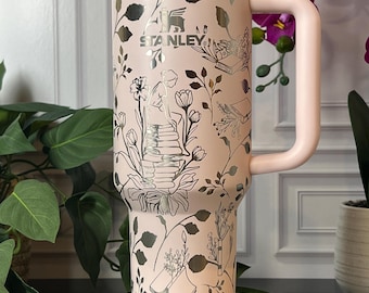 Op de afbeelding: Een roze Stanley-beker met een bloemenpatroon en het Stanley-logo. De beker heeft een handvat, een deksel en een rietje. Het bloemenpatroon is zilverkleurig en bevat bloemen, bladeren en boeken. De beker staat op een spiegeloppervlak.