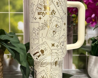 May include: Cream-coloured Stanley tumbler with gold accents and a handle. The tumbler is decorated with tarot card illustrations and the Stanley logo. It has a straw and a lid. The background includes plants and a mirror.