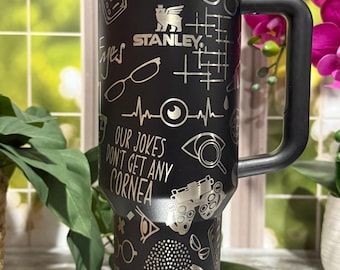 May include: Black Stanley tumbler with a handle, featuring etched designs of eyeglasses, eye charts, and the text "OUR JOKES DON'T GET ANY CORNEA" and "FIX OUR PROBLEMS." The tumbler has a black lid and straw.