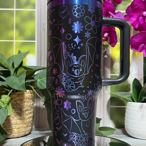 Könnte beinhalten: Schwarzer Reisebecher mit Henkel, verziert mit einem Fledermaus- und Blumendesign in schillerndem Lila und Blau. Der Becher hat einen schwarzen Deckel und Henkel. Das Design umfasst Fledermäuse, Blumen, Sterne und andere dekorative Elemente.
