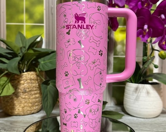 May include: Pink Stanley tumbler with a handle, straw, and a white lid. The tumbler is decorated with a pattern of dog faces, bones, and paw prints. The Stanley logo is visible. The tumbler is sitting on a reflective surface.