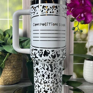 May include: A white and silver insulated tumbler with a handle, featuring a black and white spotted pattern. The tumbler has the words "Composition Book" printed on it. The tumbler is sitting on a mirrored surface.