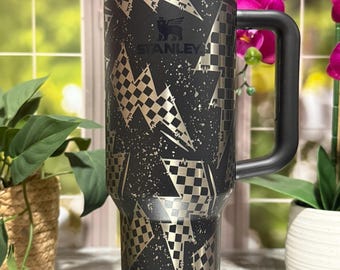 May include: A black Stanley tumbler with a handle, featuring a gold and black checkered and lightning bolt design. The tumbler has a black lid and straw. The Stanley logo is visible on the front.