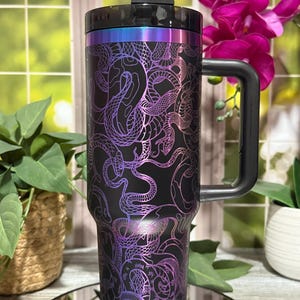 Pode incluir: Caneca de viagem preta com alça, com um design de cobra roxa e azul. A caneca tem uma tampa e alça pretas, com uma faixa metálica na parte superior. O design é um padrão repetido de cobras.