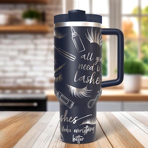 May include: A navy blue travel mug with a handle and a lid. The mug is decorated with silver illustrations of eyelashes, mascara wands, and the text "all you need is lashes" and "lashes make everything better".