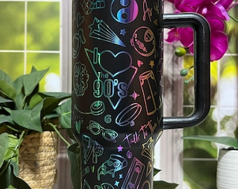 May include: Black travel mug with a handle, featuring a rainbow iridescent top and a design of 90s-themed icons. The mug includes the text "The 90's", "Be Kind", and "Chill Pill".